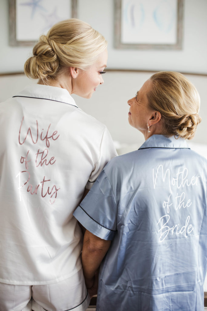 bride and mother with back turned to camera smiling before intimate beach wedding wearing matching pajamas