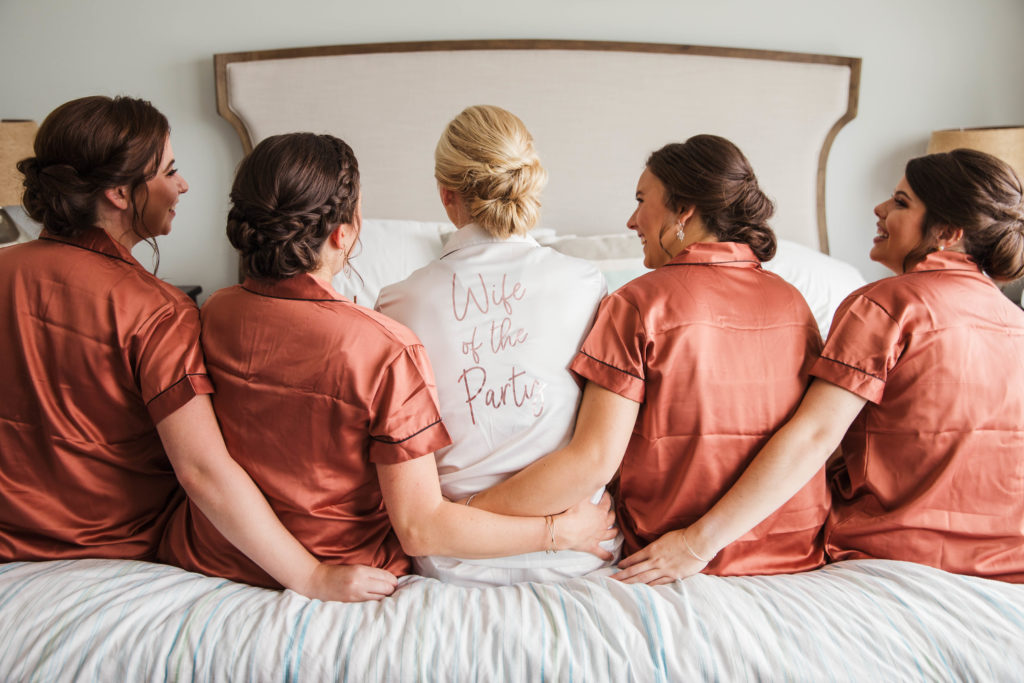 bride and friends sitting on bed in pajamas celebrating and laughing on wedding day