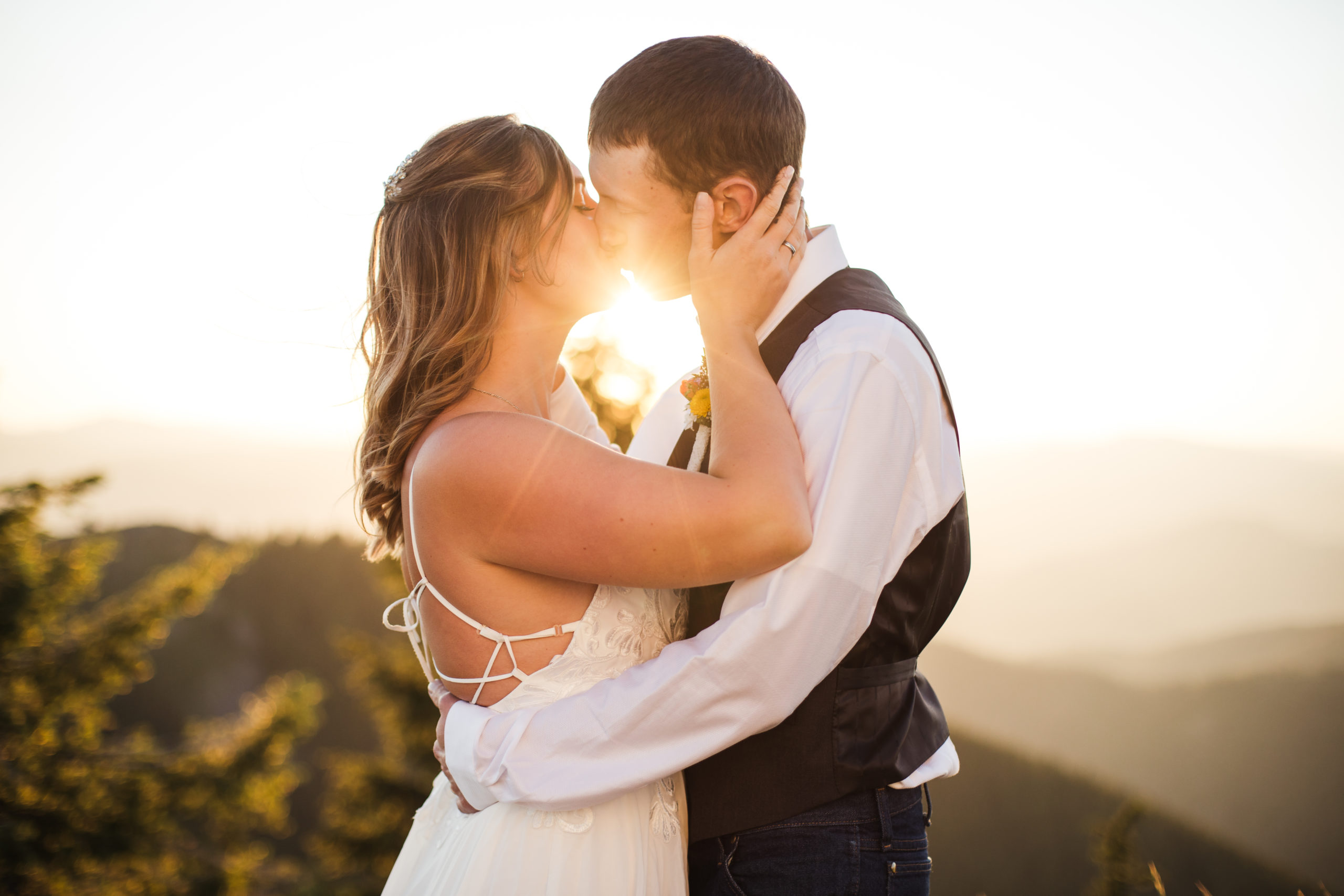 bride and groom kissing at sunset while bride wears a strappy back wedding gown at mountain top elopement