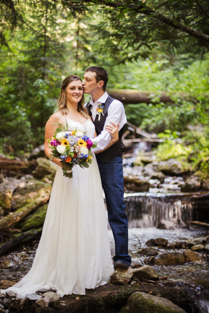 groom holds bride near waterfall while bride holds bridal bouquet with red flowers and sunflowers at mountain top elopement