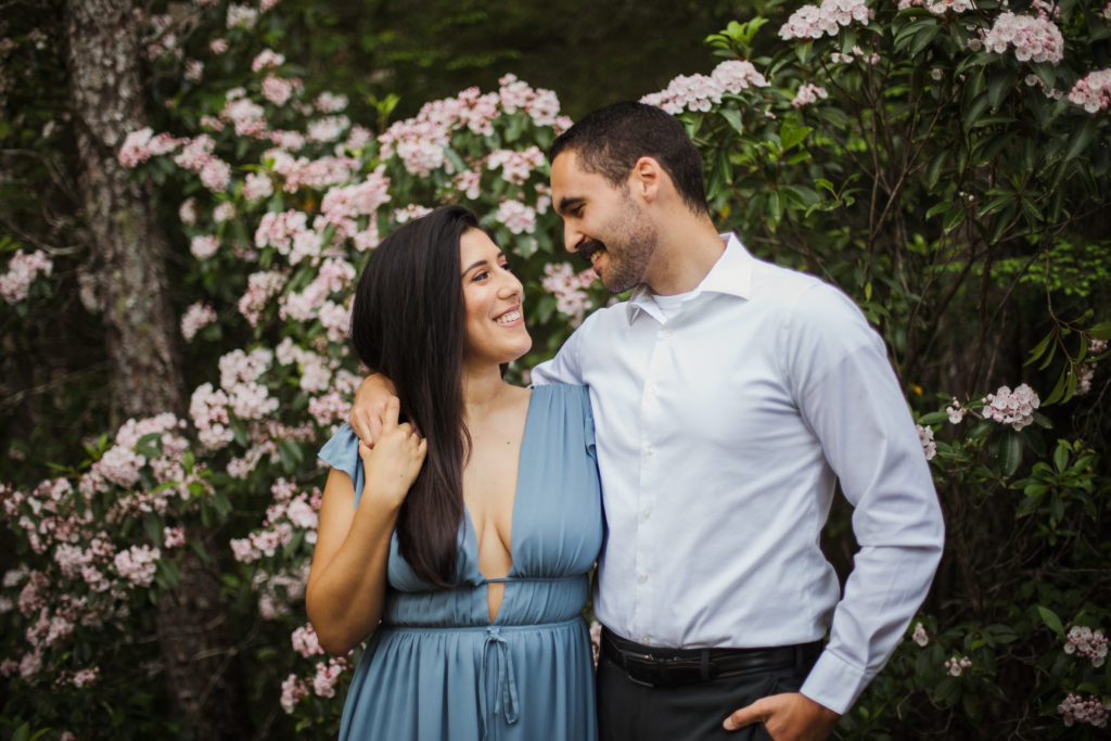 man with arm around woman's shoulder in front of blossoms during engagement session